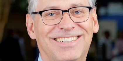 Leende man med glasögon och grått hår i blå kavaj, porträtt mot suddig bakgrund.