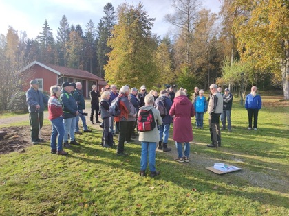 En grupp människor står utomhus på gräs, lyssnar på en guide vid en röd stuga i höstlandskap.