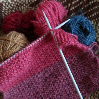 Närbild på stickning med rosa garn, stickor och garnnystan i olika färger.