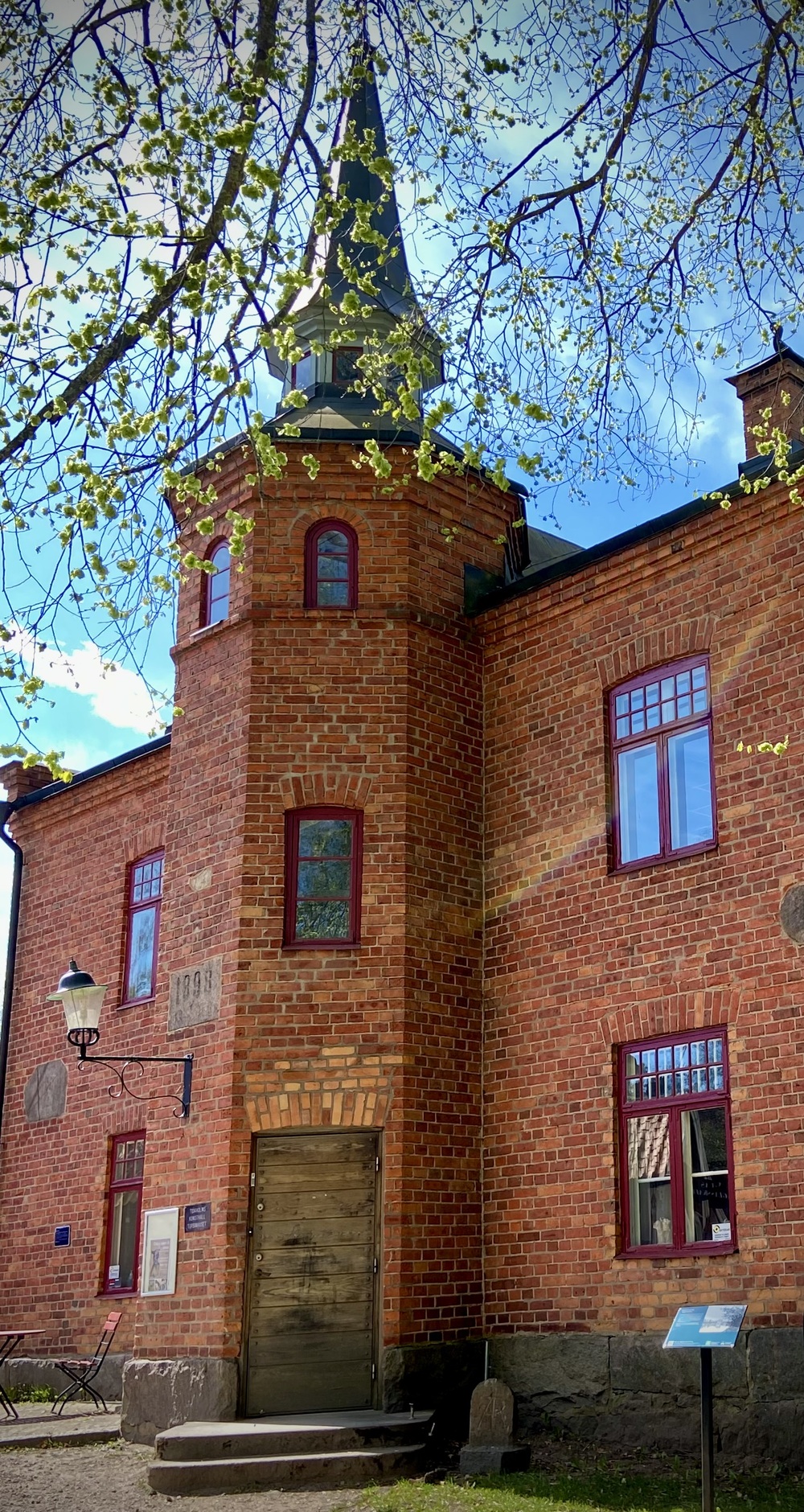 Röd tegelbyggnad med torn och spetsigt tak, omgiven av grönskande träd under blå himmel.