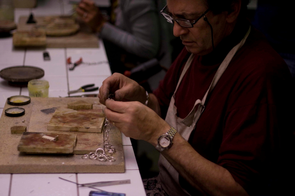 En person med glasögon och förkläde arbetar med smyckestillverkning vid ett bord med verktyg och material.