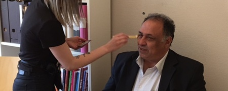 En kvinna sminkar en man som sitter vid ett bord i ett kontorsrum med bokhyllor i bakgrunden.