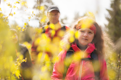 En flicka i rosa jacka och pannband vandrar i skogen med två vuxna bakom sig, omgiven av gula löv.