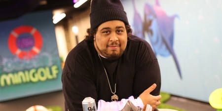 Man med mössa lutar sig mot ett bord med sprayburk och ansiktsmask i en inomhus minigolfmiljö.