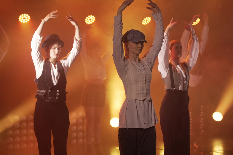 Flera personer dansar på scen i scenkläder och hattar, med armarna uppsträckta i balettposition.