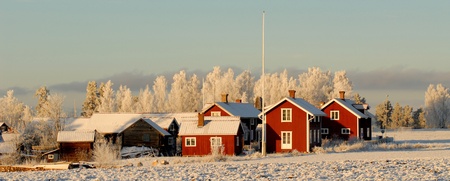 Röda trähus på snötäckt landsbygd med frostiga träd i bakgrunden under klar himmel.