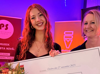 Två personer håller en stor prischeck och en blombukett på en scen med färgglad belysning.