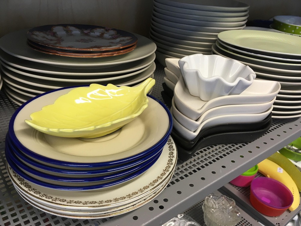 Olika staplade tallrikar och fat i olika former och färger på en hylla i ett kök eller butik.