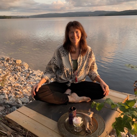Kvinna sitter i meditationsställning på en brygga vid en sjö i solnedgången.