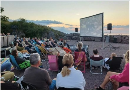 En grupp människor tittar på film utomhus vid havet i solnedgången, sittande på stolar och filtar.