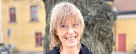 Leende kvinna med glasögon står utomhus framför ett stort träd och gula byggnader i bakgrunden.