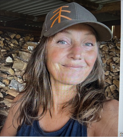 Kvinna med långt hår och keps tar en selfie framför en stapel ved inomhus.