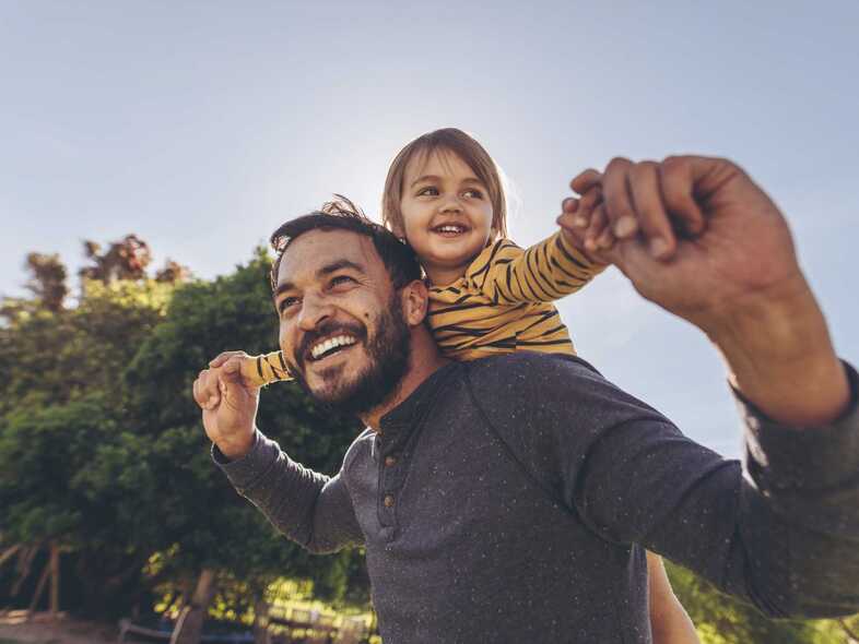En leende man med skägg har ett litet barn på ryggen och håller i dess händer.