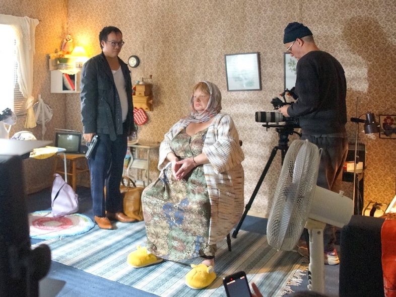 Scen vid filminspelning. En kvinna sitter på en stol mitt i ett rum. Bredvid henne står en kameraman och hennes motskådespelare, en man i kostym med en pistol i handen.