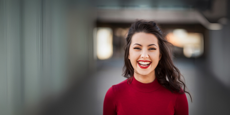 Leende person med långt mörkt hår och röd tröja står utomhus med suddig bakgrund.