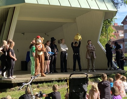 En grupp människor, några utklädda, står på en utomhusscen inför en sittande publik i solsken.