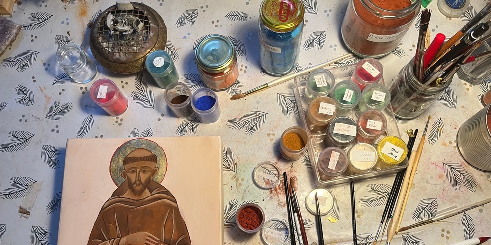 Ett bord med konstnärsmaterial, penslar, färgburkar och en ikonmålning av en helig man.