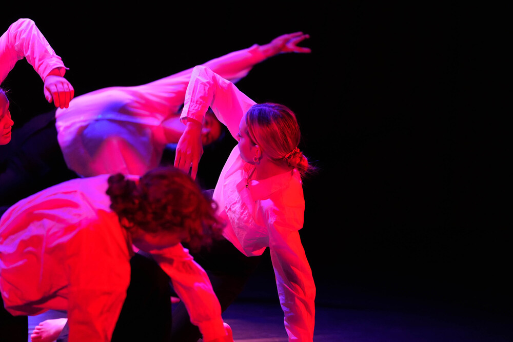 Tre personer dansar i dramatiskt röd belysning på en mörk scen, klädda i vita skjortor.