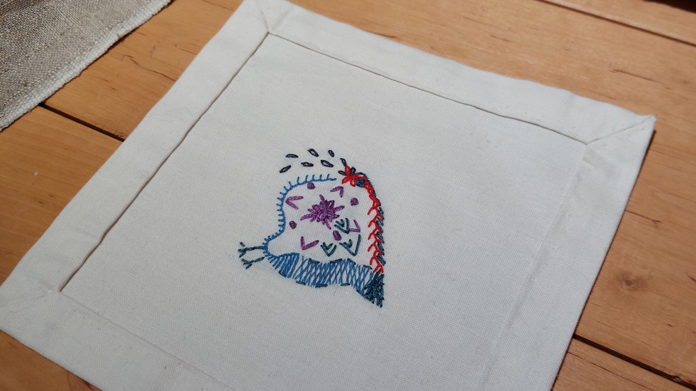 En tygbit med broderat mönster i olika färger ligger på ett trägolv.