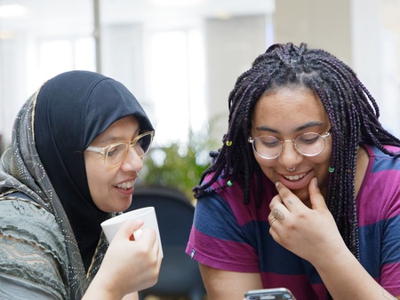 Två unga kvinnor sitter tillsammans, ler och tittar på en mobiltelefon.