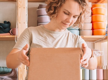 En person packar en kartong i en ljus och organiserad verkstad med hyllor fyllda av keramik och förvaringslådor.