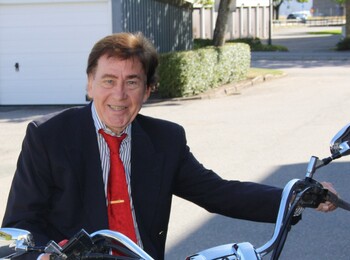 Äldre man i kostym och röd slips sitter på en motorcykel utomhus vid en gata.