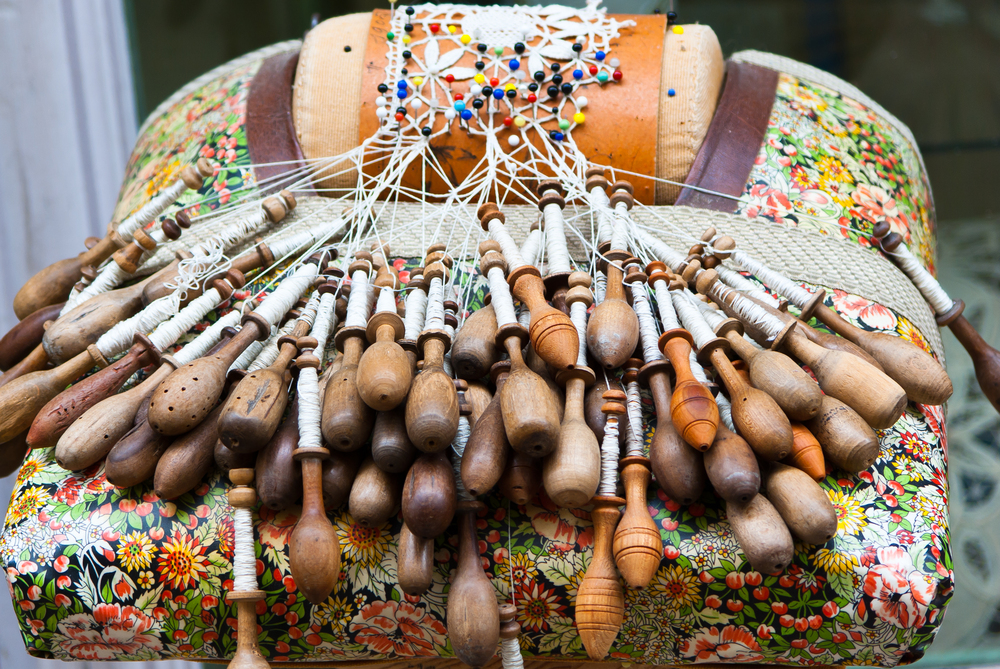 Närbild av en spetskudde med många träspolar och pågående knypplingsarbete.