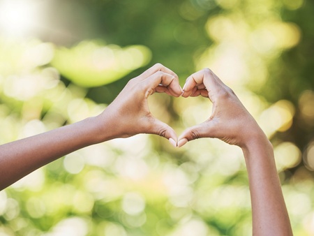 Två händer formar ett hjärta mot en suddig, grön bakgrund utomhus.