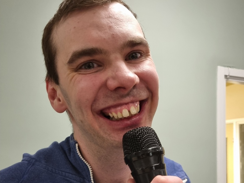 Leende man i blå tröja håller en mikrofon och tittar in i kameran.