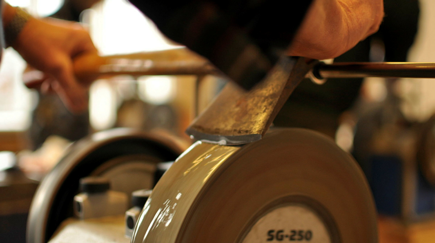 En person slipar ett metallverktyg mot en roterande slipsten i en verkstadsmiljö.