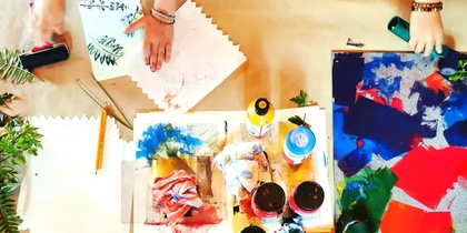Två personer arbetar med färgglada konstmaterial, papper och färg på ett bord.
