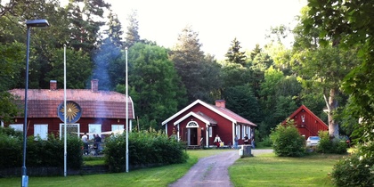 En grusväg leder till tre röda hus omgivna av grönska och träd, med flaggstänger och en grillplats.