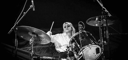 Kvinna spelar trummor på en scen, svartvit bild med mikrofoner och trumset i fokus.