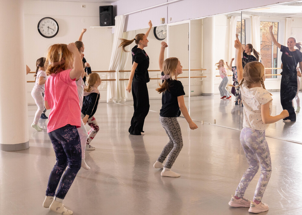Barn och en vuxen dansar tillsammans i en ljus dansstudio med spegelvägg och klocka på väggen.