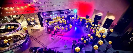 Vy ovanifrån av ett evenemang inomhus med många människor, scen, färgglad belysning och text på väggen.