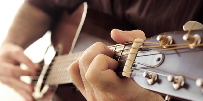 Närbild av en person som spelar akustisk gitarr, fokus på vänster hand på gitarrens greppbräda.