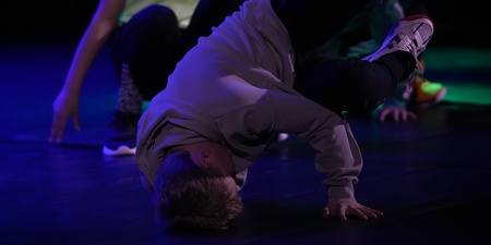 En person utför ett breakdance-move på golvet med stöd av ena handen, andra personer i bakgrunden.