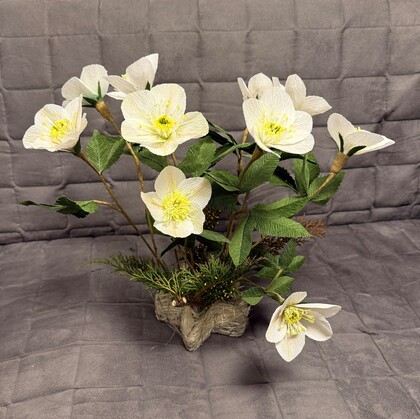 En bukett vita konstgjorda blommor med gröna blad i en dekorativ bas, placerad mot en grå vadderad bakgrund.