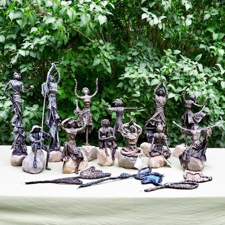 Flera små skulpturer av dansande människor står på ett bord utomhus med grönska i bakgrunden.
