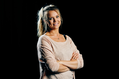Kvinna med ljust hår i hästsvans står med armarna i kors och ler mot kameran, svart bakgrund.