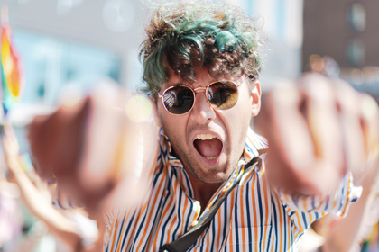 En person med solglasögon och randig skjorta sträcker ut knytnävarna mot kameran och ropar utomhus.