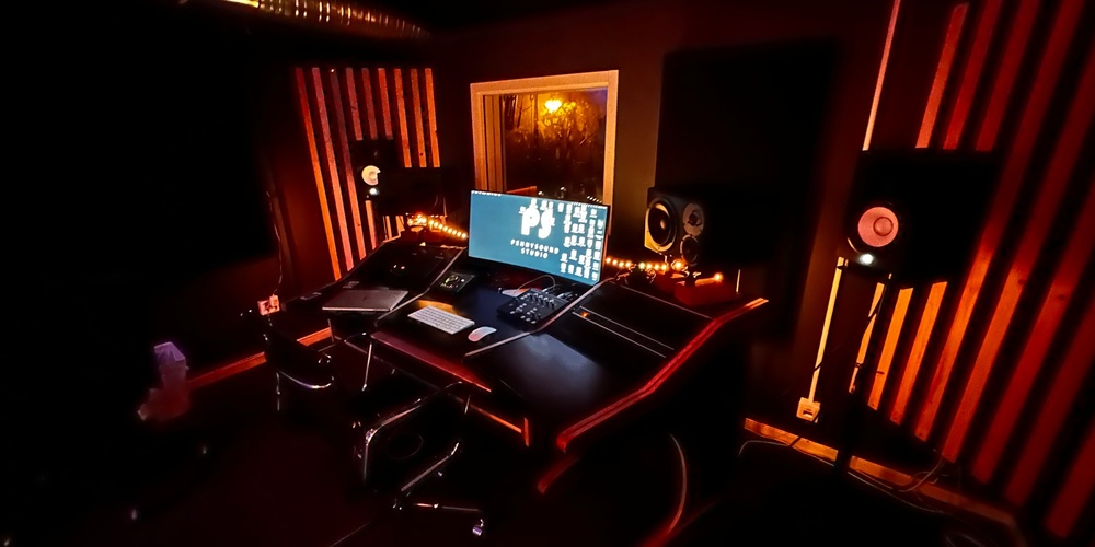 Ett mörkt inspelningsstudio med orange belysning, dator, högtalare och ljudutrustning på ett skrivbord.