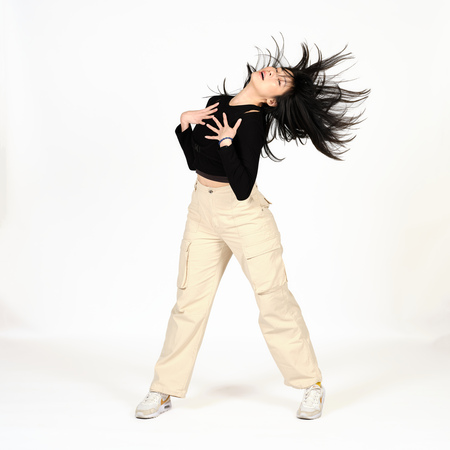 En person med långt hår dansar energiskt i ljusa byxor och svart tröja mot vit bakgrund.