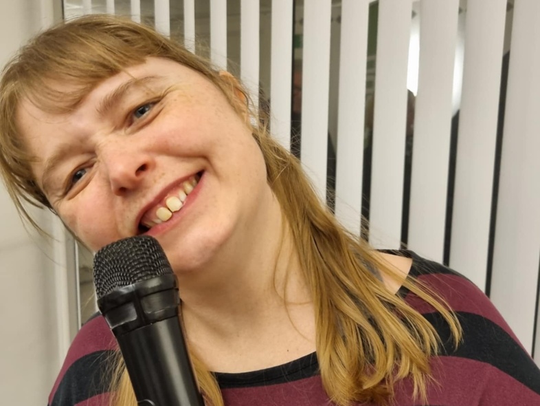 Leende kvinna med blont hår håller en mikrofon och står framför vita persienner.