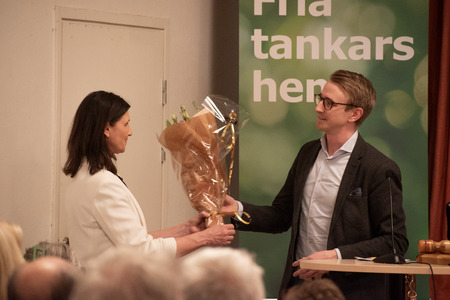 En kvinna tar emot en blombukett från en man under ett evenemang med publik. En grön skylt syns i bakgrunden.