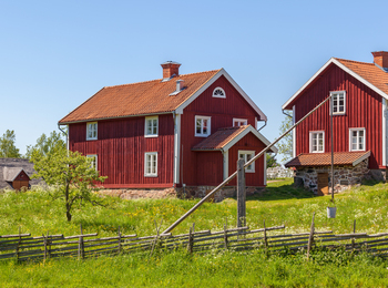 Två röda trähus med vita knutar på landet, omgivna av gräs, staket och blå himmel.
