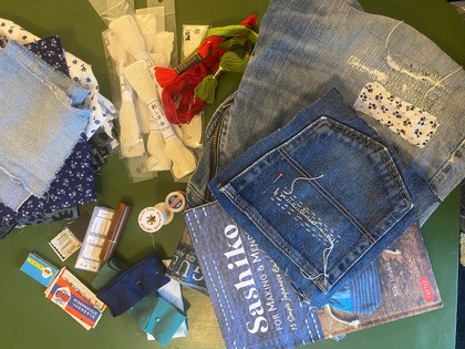 Material för sashiko-broderi, inklusive tygbitar, tråd, nålar, broderade jeans och en instruktionsbok på ett grönt bord.