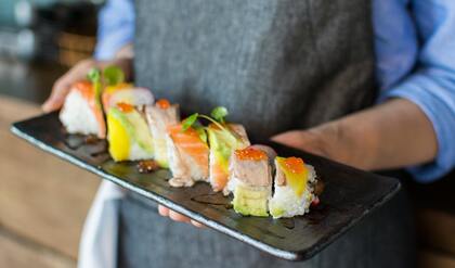 En person i förkläde håller en rektangulär tallrik med färgglada sushibitar.