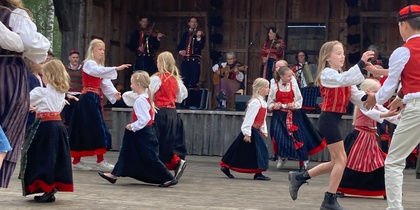 Barn och vuxna i folkdräkt dansar på en utomhusscen med musiker i bakgrunden.