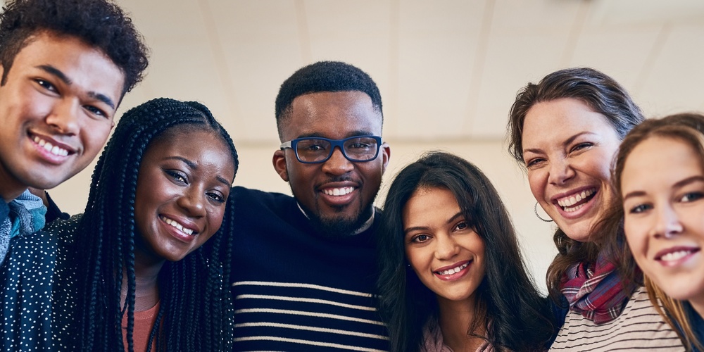 Sex unga vuxna står tätt tillsammans och ler mot kameran i ett ljust rum.